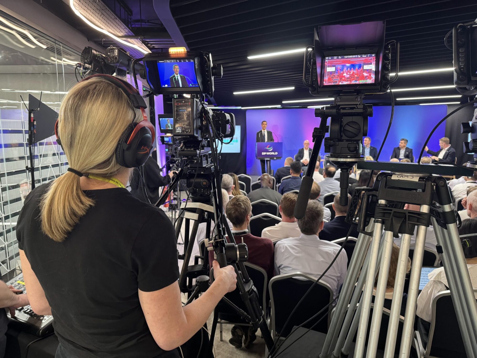 A woman with headphones operates a camera at a press conference, capturing a speaker at a podium. The audience, consisting of seated individuals, is visible in the background, along with several cameras and broadcast equipment.