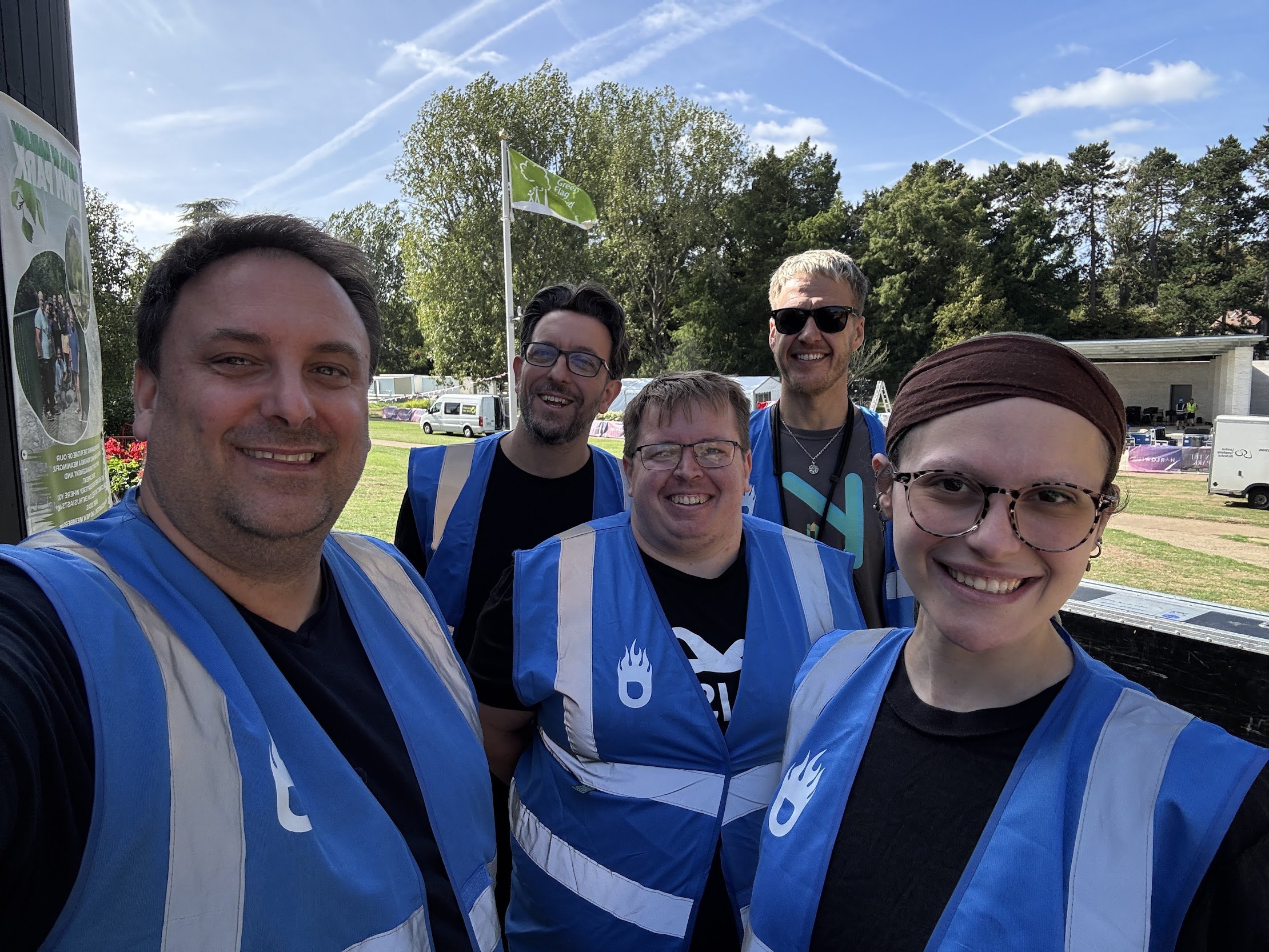 Five smiling oXyFire crew members wearing blue production vests at an outdoor event in Harlow
