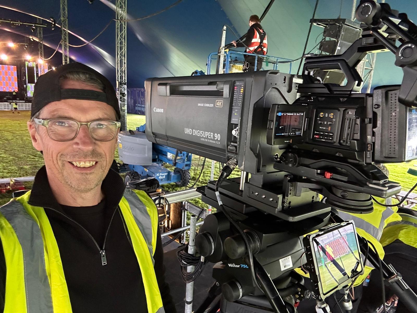 Smiling camera operator beside a broadcast camera at Reminisce festival